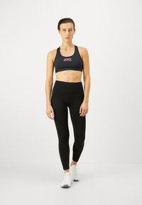 Sujetador deportivo negro con logo "NIKE" en rosa, combinado con leggings negros de cintura alta. Modelo llevando zapatillas de running blancas; fondo minimalista.