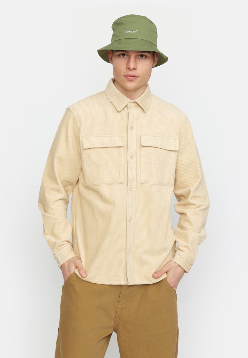 Beige corduroy overhemd met twee klepzakken, knoopsluiting en kraag; gecombineerd met kaki broek en een groene emmerhoed met logo.