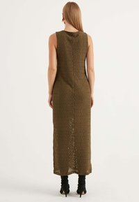 Abito maxi senza maniche di colore verde oliva, realizzato in materiale a maglia testurizzato con motivo a zigzag. Presenta una fessura a metà schiena e stivaletti neri con tacco alti fino alla caviglia.