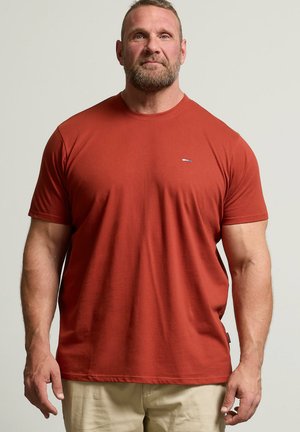 Uomo muscoloso con barba che indossa una maglietta a maniche corte color ruggine e pantaloni beige, in piedi davanti a uno sfondo chiaro uniforme.