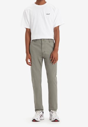 Mann trägt weißes Levi's T-Shirt, graue Hose mit geradem Bein und weiße Sportschuhe, steht vor weißem Hintergrund.