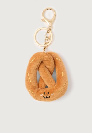 Porte-clés en forme de bretzel en peluche avec un visage souriant et un fermoir de couleur dorée sur fond blanc uni.