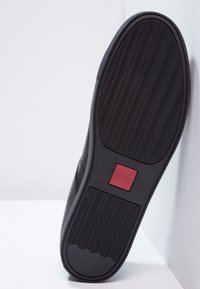 Schwarze Sneaker-Sohle aus strukturiertem Gummi mit einem roten, rechteckigen Hugo-Logo. Das Design umfasst wellenförmige Profilmuster für besseren Halt.