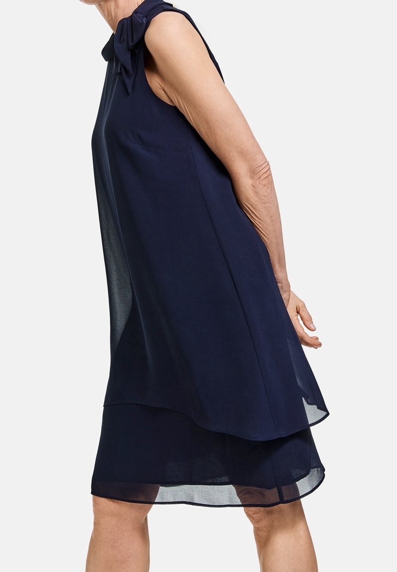 Femme senior portant une robe bleu marine sans manches à couches avec un détail nœud sur l'épaule, debout devant un fond uni.