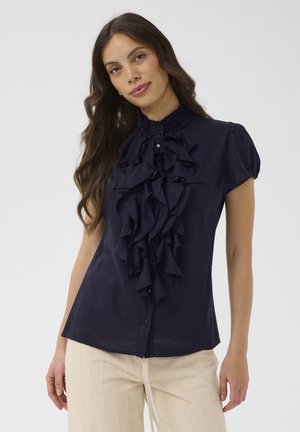 Femeie cu păr lung și întunecat poartă o bluză bleumarin cu volane, mâneci scurte bufante și nasturi în față, asortată cu pantaloni bej deschis.