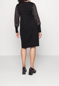 Femme portant une robe noire mi-longue avec des manches longues transparentes noires ornées de pois dorés, associée à des chaussures noires à talons carrés.