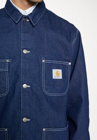 Denim arbetsjacka i mörkblått. Har silverknappar, två framfickor med vit söm och en Carhartt-logotagg på vänstra fickan.