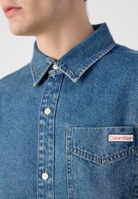 Camisa de denim en azul medio, con collar de punta, botones blancos y un bolsillo en el pecho con una etiqueta de Calvin Klein roja y blanca.