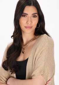 Cardigan beige a maglia con maniche corte, collana a catena d'oro a più strati con pendente, su uno sfondo neutro. Top scuro sotto il cardigan.