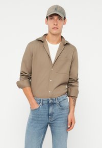 Cămașă lungă bej deschis cu mâneci lungi, cu nasturi în față și buzunar; purtată peste un tricou alb. Asortată cu blugi ușor albaștri și o șapcă gri.