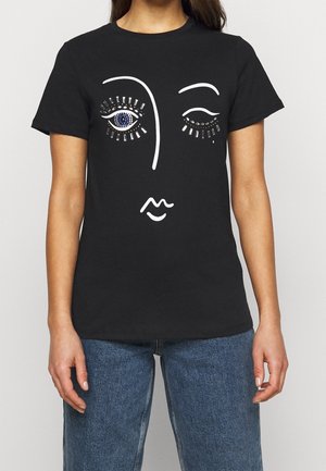 Black t-shirt with abstract white face outline featuring one open eye with blue and silver embellishments and one winking eye, worn with blue jeans.