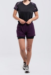 Černé sportovní tričko s fialovým květinovým motivem na ramenou, doplněné tmavě fialovými šortkami přes černé kompresní šortky a černými teniskami.