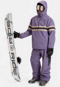 Lilla snowboardjakke med beige og sorte striber, matchet med lilla snowboardbukser. Personen holder et hvidt snowboard med sorte detaljer.