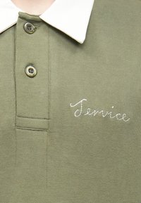 Polo de color verde oliva con cuello blanco, con dos botones y texto bordado "Temrice" en cursiva blanca en la parte delantera.