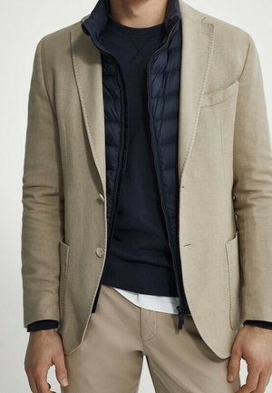 Homme portant un blazer beige sur un gilet matelassé zippé bleu marine et un pull bleu marine avec un pantalon kaki clair, sur un fond clair uni.