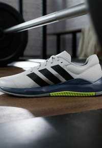 Zapatilla deportiva blanca con rayas negras, parte superior texturizada, suela azul y detalle en verde neón. Diseñada para el rendimiento y la estabilidad.