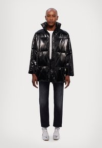 Chaqueta tipo puffer negra con un acabado brillante, corte oversized, cuello alto y detalles en botones. Combinada con jeans oscuros y zapatos plateados.