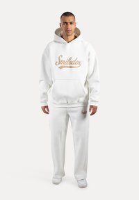 Conjunto de sudadera blanca y pantalones de chándal con un bolsillo frontal y el logo "Smilodox" en marrón claro. Textura suave, corte relajado y diseño clásico.