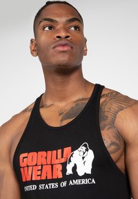 Črn top brez rokavov s rdečim in belim logotipom "GORILLA WEAR" ter grafičnim oblikovanjem. Material se zdi gladek, ima zaobljen izrez.