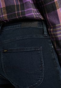 Jean en denim foncé avec une coupe ajustée, un léger éclat, des poches arrière et une étiquette logo Lee bien mise en avant à la taille.