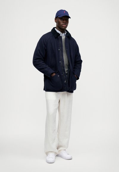 Homem a usar um casaco azul-marinho sobre um suéter cinza e uma camisa xadrez, calças brancas, ténis brancos e um boné azul-marinho, de pé com as mãos nos bolsos.