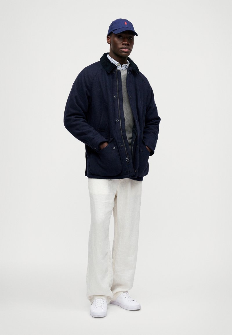 Homme portant une veste bleu marine par-dessus un pull gris et une chemise à carreaux, un pantalon blanc, des sneakers blanches et une casquette de baseball bleu marine, se tenant avec les mains dans les poches.