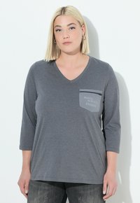 Blonde vrouw in een grijze V-hals T-shirt met 3/4 mouwen en een ritssluiting op de borstzak met de tekst "ROCK & ROLL INSIDE" tegen een witte achtergrond.
