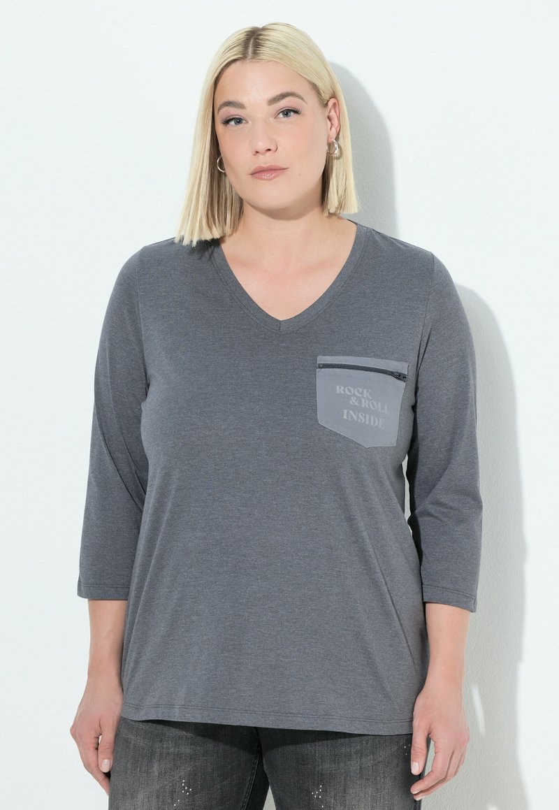 Blonde vrouw in een grijze V-hals T-shirt met 3/4 mouwen en een ritssluiting op de borstzak met de tekst "ROCK & ROLL INSIDE" tegen een witte achtergrond.