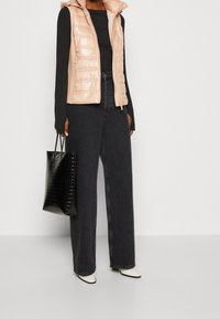 Lekko wyściełana, błyszcząca beżowa kamizelka z kapturem, noszona na czarną bluzkę z długim rękawem. Czarne, szerokie dżinsy i fakturowana czarna torba na ramię. Białe czółenka w szpic.