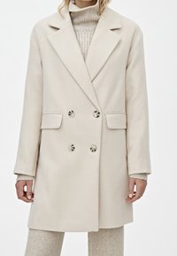 Cappotto beige doppiopetto con revers, dotato di due tasche frontali e due grandi bottoni, realizzato in un morbido tessuto testurizzato.
