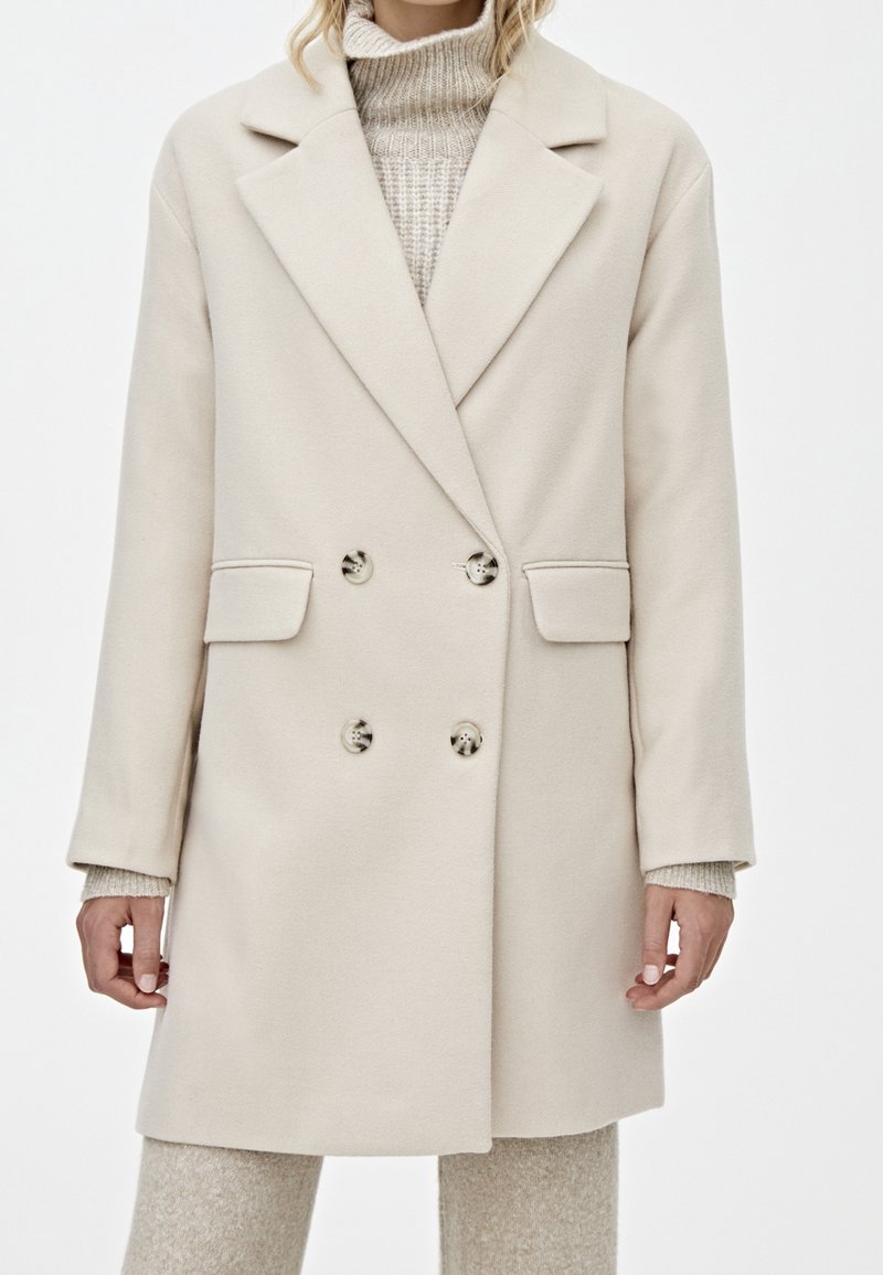 Cappotto beige doppiopetto con revers, dotato di due tasche frontali e due grandi bottoni, realizzato in un morbido tessuto testurizzato.