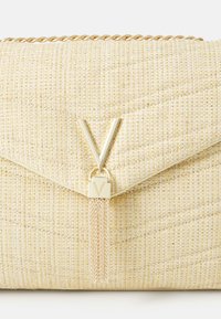 Πολυτελής τσάντα χειρός από τυλιγμένο μπεζ ύφασμα με χρυσές λεπτομέρειες, διαθέτοντας κεντρικό χρυσό κούμπωμα με το λογότυπο «V» και μια λεπτομέρεια με αλυσσίδα και φούντα.