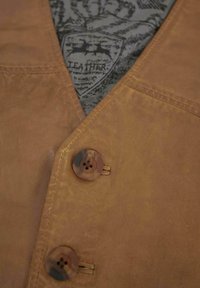 Braune Lederweste mit V-Ausschnitt, ausgestattet mit zwei runden Knöpfen und einem gemusterten grauen Futter, das ein Markenzeichen aus Leder zeigt.