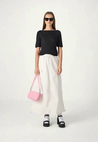 Mulher com blusa preta, saia longa branca, sandálias de plataforma pretas com meias brancas, óculos de sol pretos, segurando uma bolsa rosa.