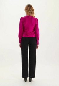 Blouse fuchsia avec des manches bouffantes, texture lisse et boutons aux poignets, associée à un pantalon noir taille haute à jambes larges et des talons.