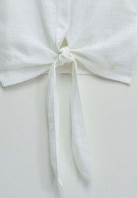 White linen fabric tied in a knot with two loose ends hanging down, featuring a small gold tag on the bottom right side.