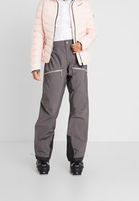 Prošívaná růžová bunda s vysokým límcem, doplněná šedými lyžařskými kalhotami s kapsami na zip a černými spodními manžetami. Outfit doplňují černé lyžařské boty.