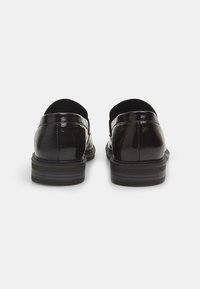 Zwarte leren loafers met een glanzende afwerking, voorzien van een ronde neus en een lage, gelaagde hak. De achterkant toont een gladde, glanzende textuur.