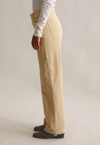 Beżowe spodnie sztruksowe o prostym kroju, z wysokim stanem, dwoma tylnymi kieszeniami i miękką fakturą. Noszone z szarymi butami na obcasie.
