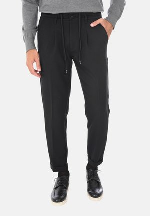 Pantaloni tapered neri con vita regolabile, parte anteriore plissettata, tasche laterali e orli arrotolati. Realizzati in tessuto liscio, abbinati a scarpe nere.