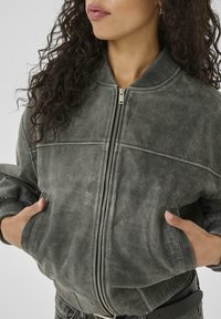 Gray suede bomber jacket with ribbed cuffs and collar, featuring a front zipper closure and side pockets. Smooth texture and tailored fit.