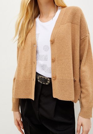 Lyshåret kvinde iført en kamelfarvet strikket cardigan over en hvid t-shirt med rhinstenprint, sorte bukser med bælte og lange kædeøreringe.
