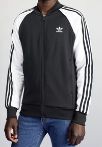 Černá bunda na zip s bílými rukávy, na každém rukávu tři černé pruhy a na hrudi logo Adidas. Vyrobena z hladké látky.