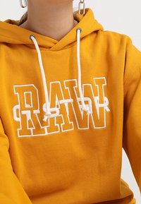 Osoba w musztardowej bluzie z kapturem, z białymi sznureczkami, z dużym białym napisem "RAW" na piersi, również nosząca srebrne prostokątne kolczyki.