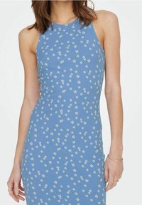 Robe sans manches bleue avec un petit motif floral blanc, portée par une femme aux cheveux bruns mi-longs.