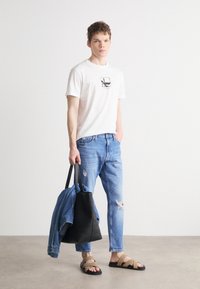 Camiseta blanca con diseño gráfico, jeans desgastados azules, bolsa de tela negra, chaqueta de mezclilla azul, sandalias de tiras marrones, fondo claro de suelo.