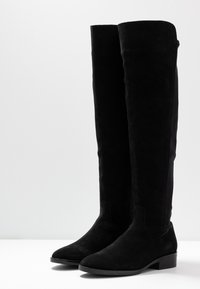 Botas negras de ante por encima de la rodilla, con un diseño elegante y un tacón plano, con una textura suave y acentos minimalistas.