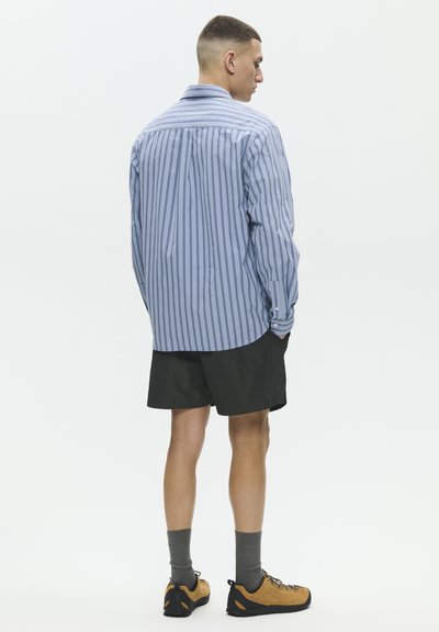 Hombre de espaldas vistiendo una camisa azul de rayas de manga larga, pantalones cortos negros, calcetas grises y zapatos de senderismo color canela sobre un fondo blanco.