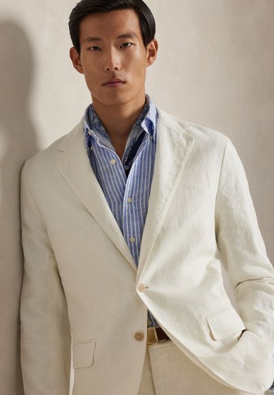 Šviesiai smėlio spalvos lino švarkas su ryškiu apykakle, derinamas su mėlynos ir baltos spalvų dryžuota marškinių tipo palaidine. Ryškūs akcentai yra raštuotas šalikas ir diržas.