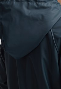 Vue de dos d'une personne portant une veste à capuche noire en tissu lisse et légèrement brillant.